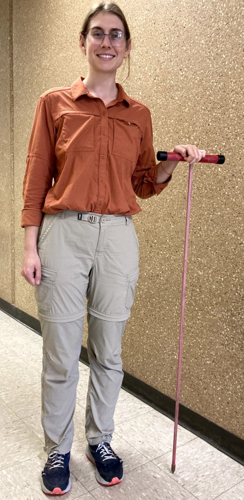 A smiling woman standing in a hallway, wearing an orange shirt and beige cargo pants, holding a pink cane.
