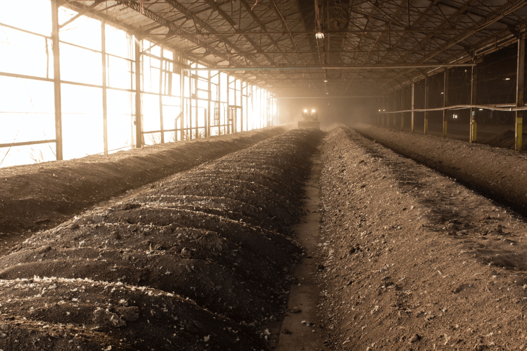 Interior of a dimly lit agricultural building with rows of soil or compost piles, and a tractor in the background dust cloud.