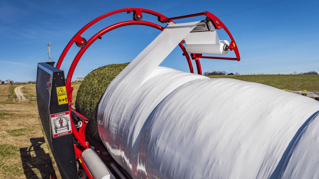 A red and black bale-wrapper machine applying white plastic wrap around a large hay bale in a field under a clear blue sky.