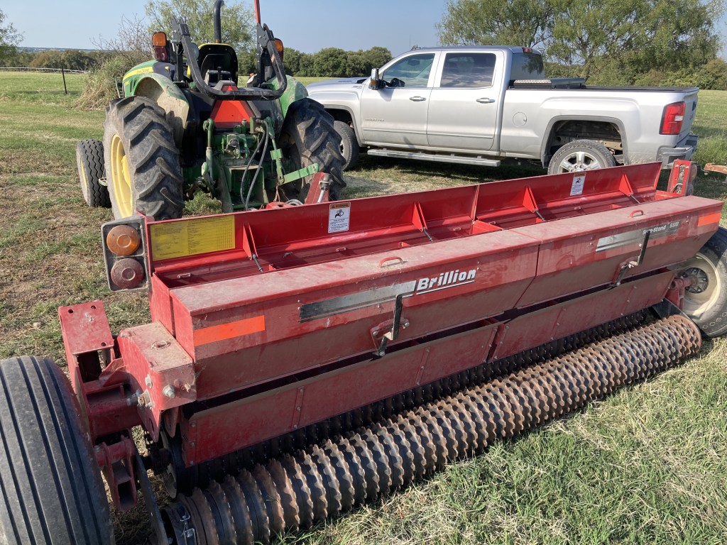 A red Brillion seed drill parked on a grassy field, with a green tractor and a silver pickup truck in the background.